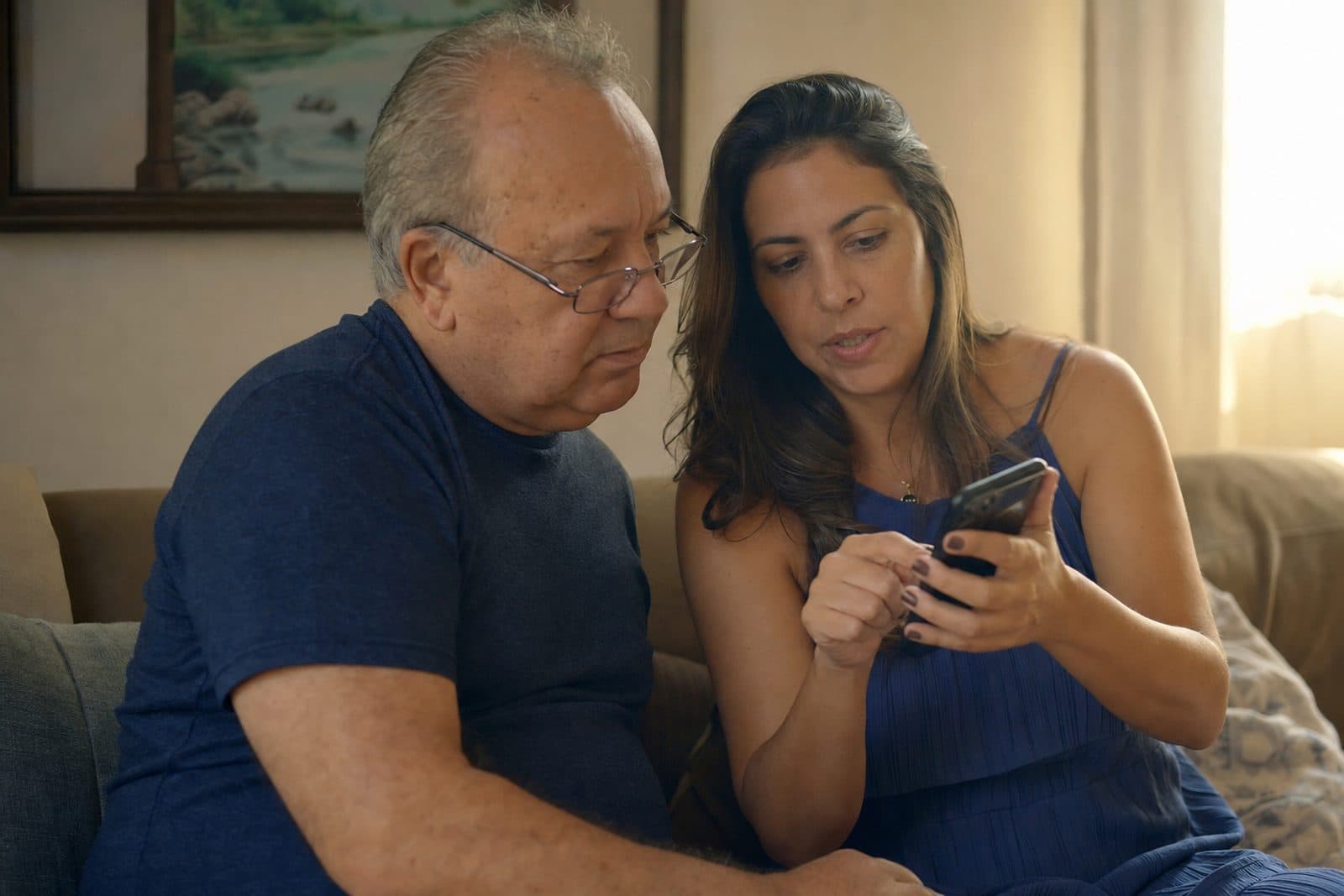 Filha mostra a tela do celular para o pai idoso sentado ao lado no sofá